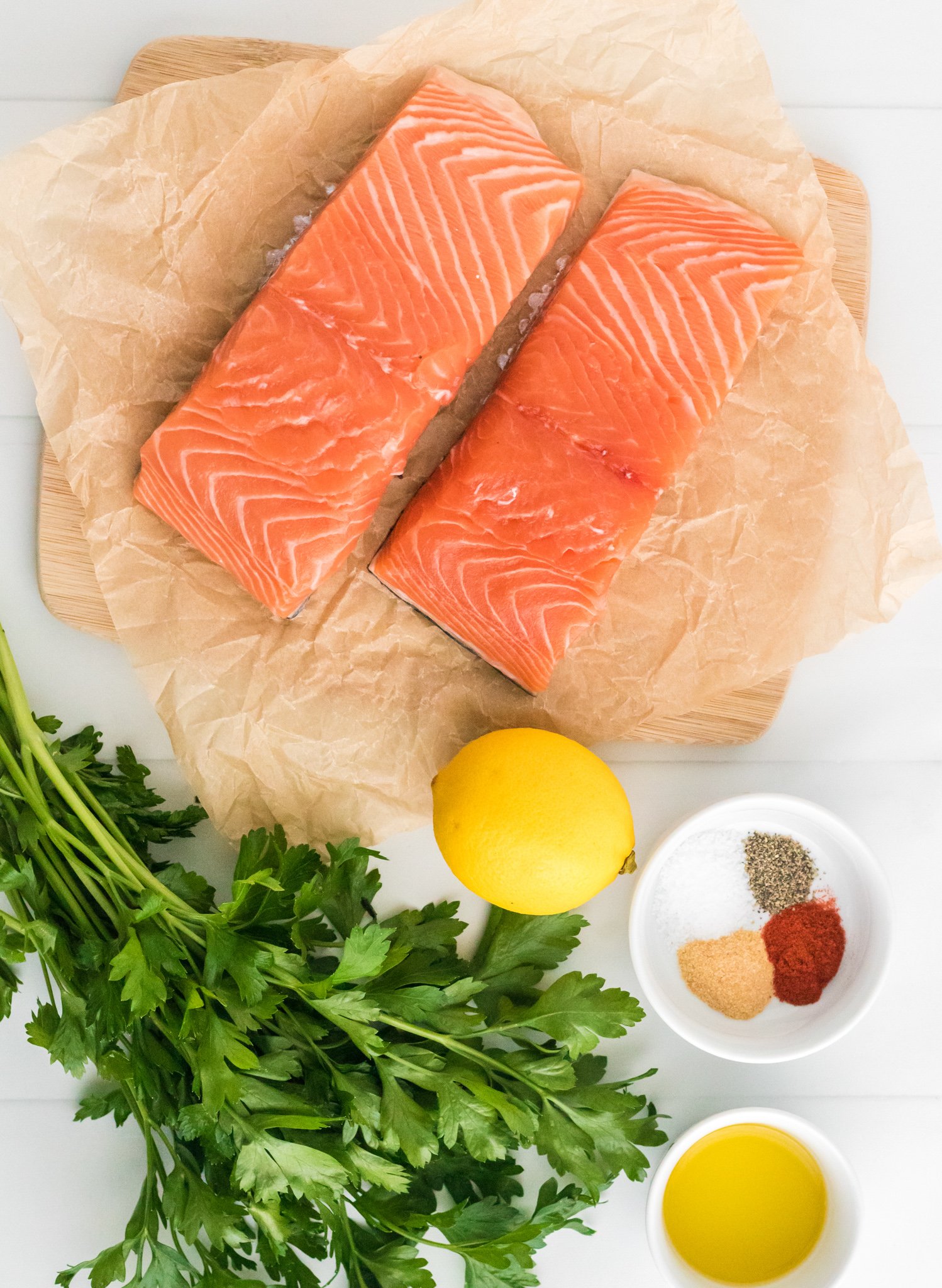 Two raw salmon fillets on parchment paper atop a wooden board. A bunch of fresh parsley, a lemon, and a small bowl containing spices, salt, and pepper are beside a bowl of olive oil on a white surface.