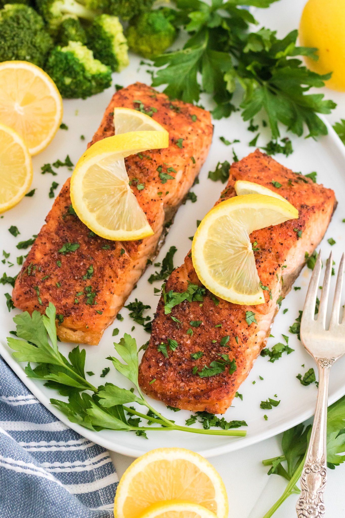 Two seasoned salmon fillets on a white plate, each garnished with a lemon slice and chopped herbs. Surrounding the salmon are sprigs of parsley and lemon wedges. A fork and broccoli are visible next to the plate.