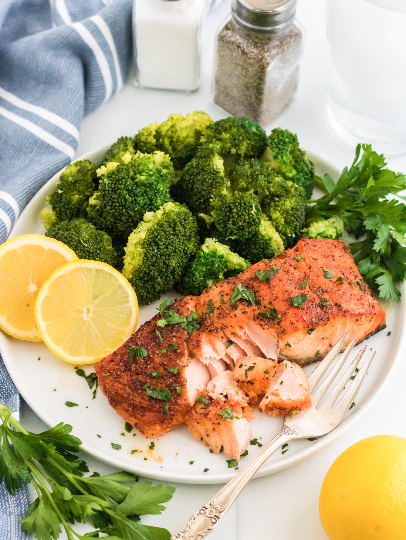 A plate with roasted salmon garnished with herbs, steamed broccoli, and lemon slices. A fork flakes the salmon. Salt and pepper shakers, a glass of water, and a striped cloth are in the background. Fresh parsley and a lemon are nearby.