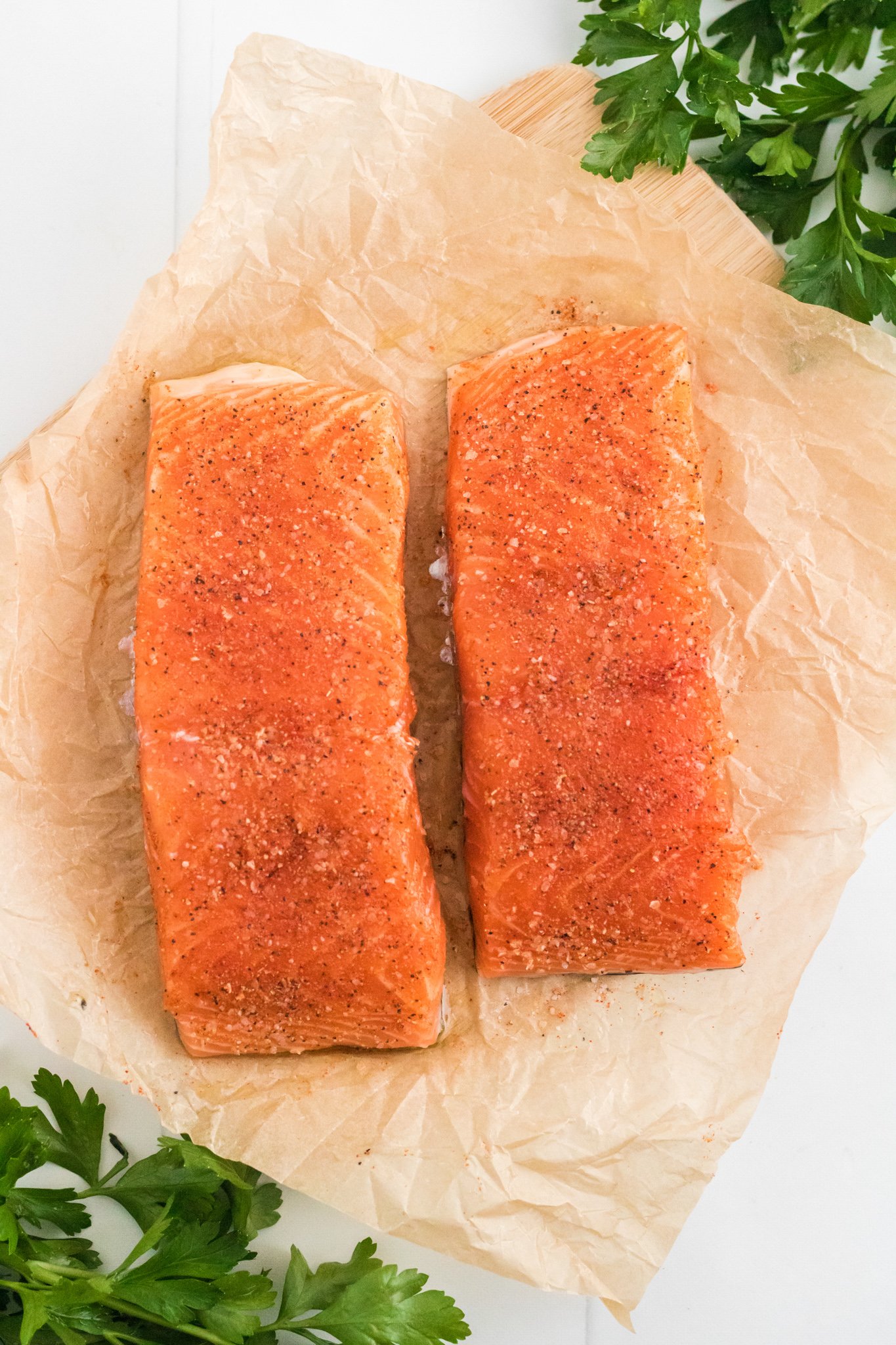 Two raw salmon fillets seasoned with spices rest on crinkled parchment paper. Fresh parsley leaves are scattered around, adding a touch of green to the image. The setup is on a light-colored surface.