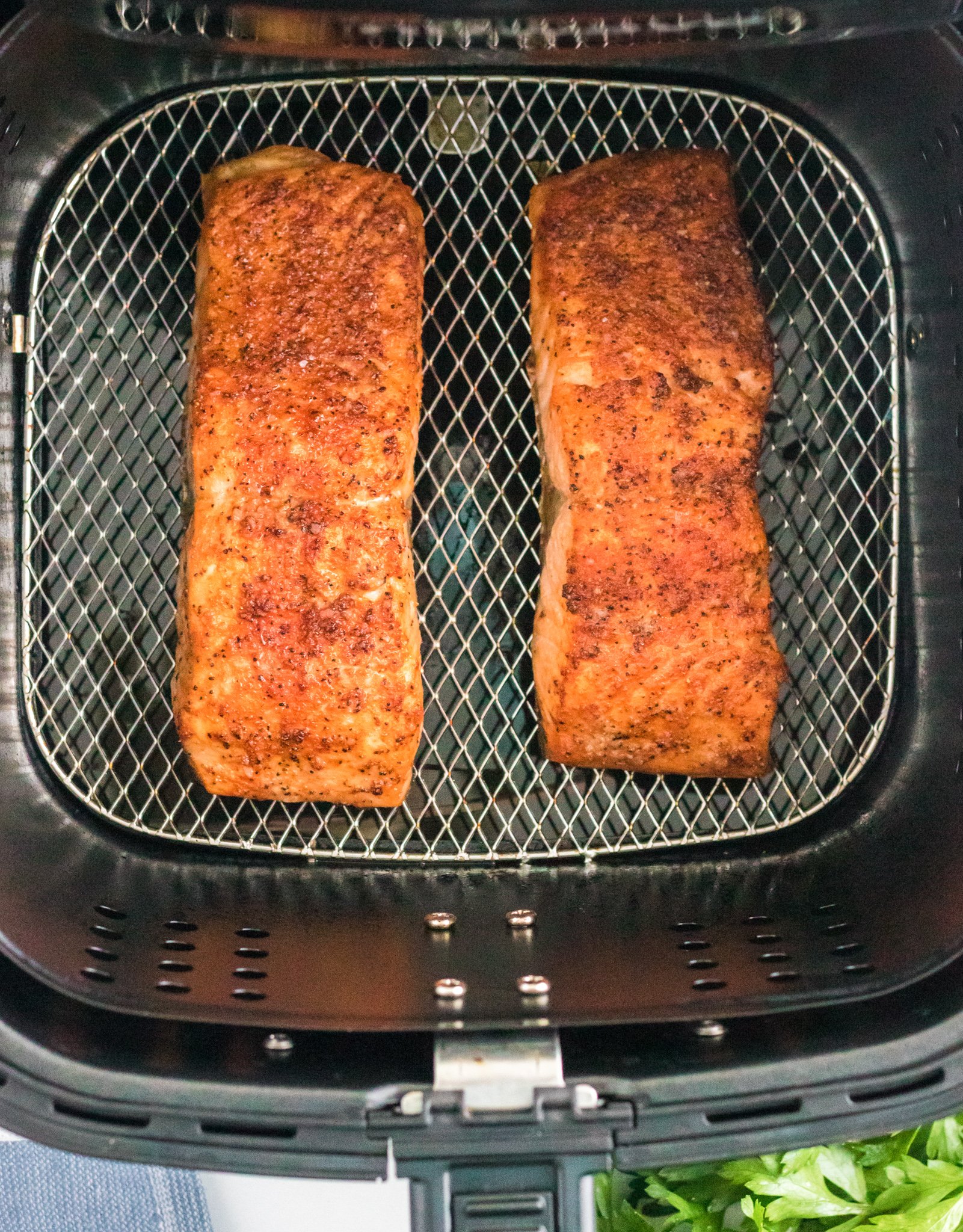 Two seasoned salmon fillets are cooking in an air fryer basket. The fillets are golden brown with a crispy texture. Fresh herbs are visible in the bottom right corner.