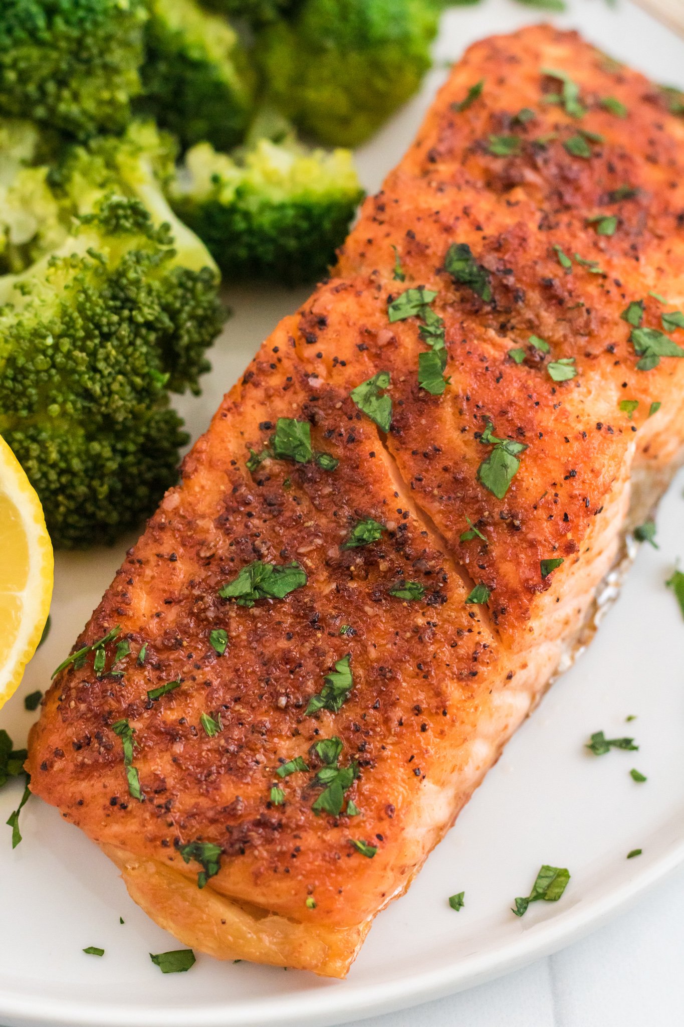 Grilled salmon fillet seasoned with herbs and pepper, garnished with chopped parsley, is served on a white plate alongside vibrant green broccoli florets and a lemon wedge.