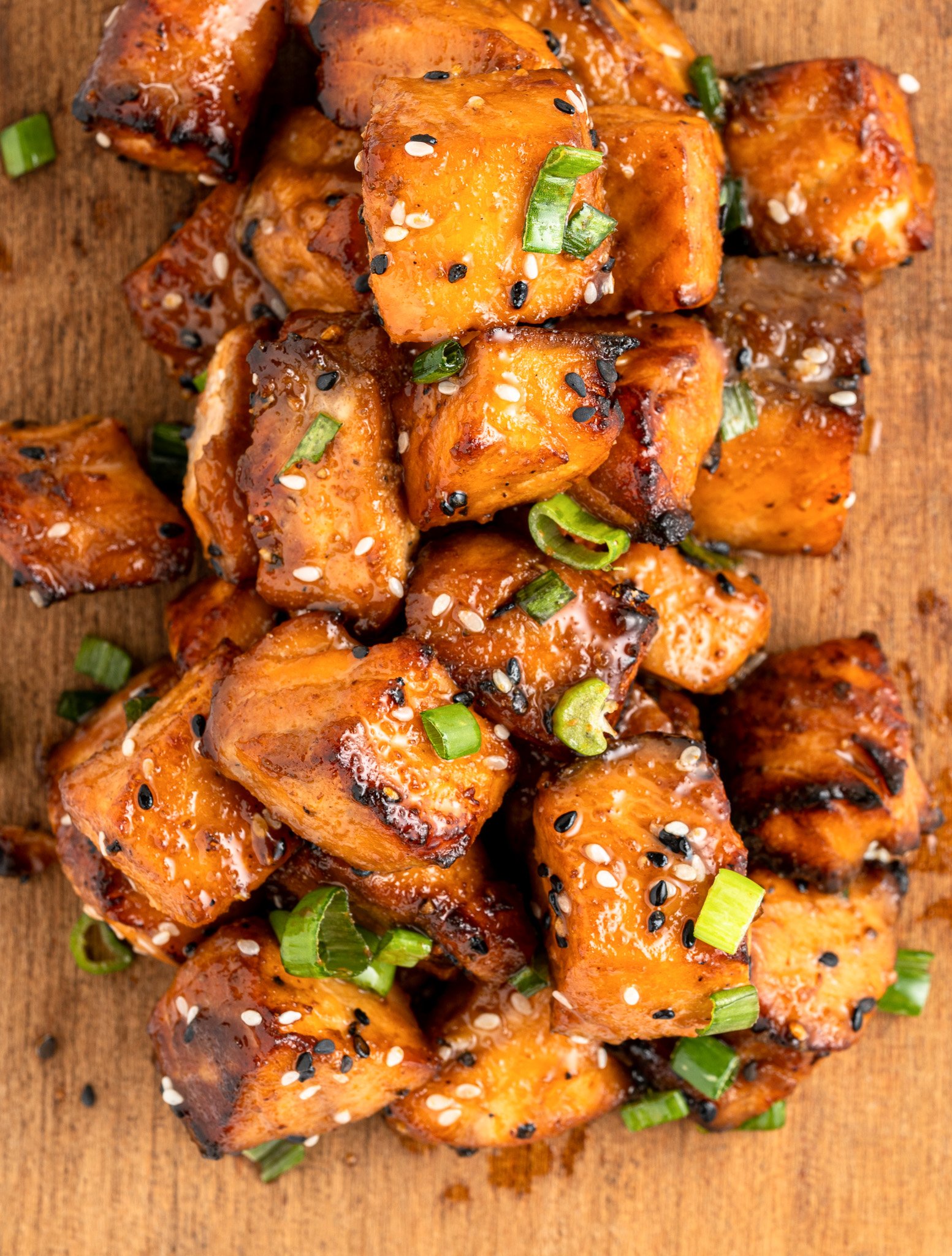 A pile of glazed, air fryer salmon bites topped with sesame seeds and sliced green onions on a wooden surface. The salmon has a caramelized appearance, indicating its well-cooked and flavorful.