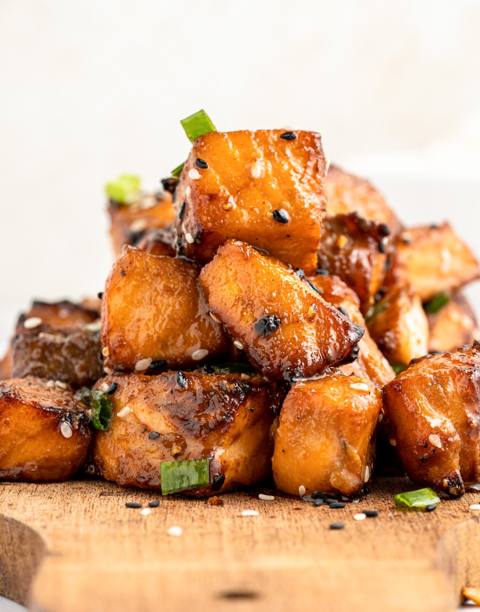 A stack of air fryer salmon bites garnished with sesame seeds and green onions on a wooden board. The pieces are golden-brown, highlighting their caramelized surface.