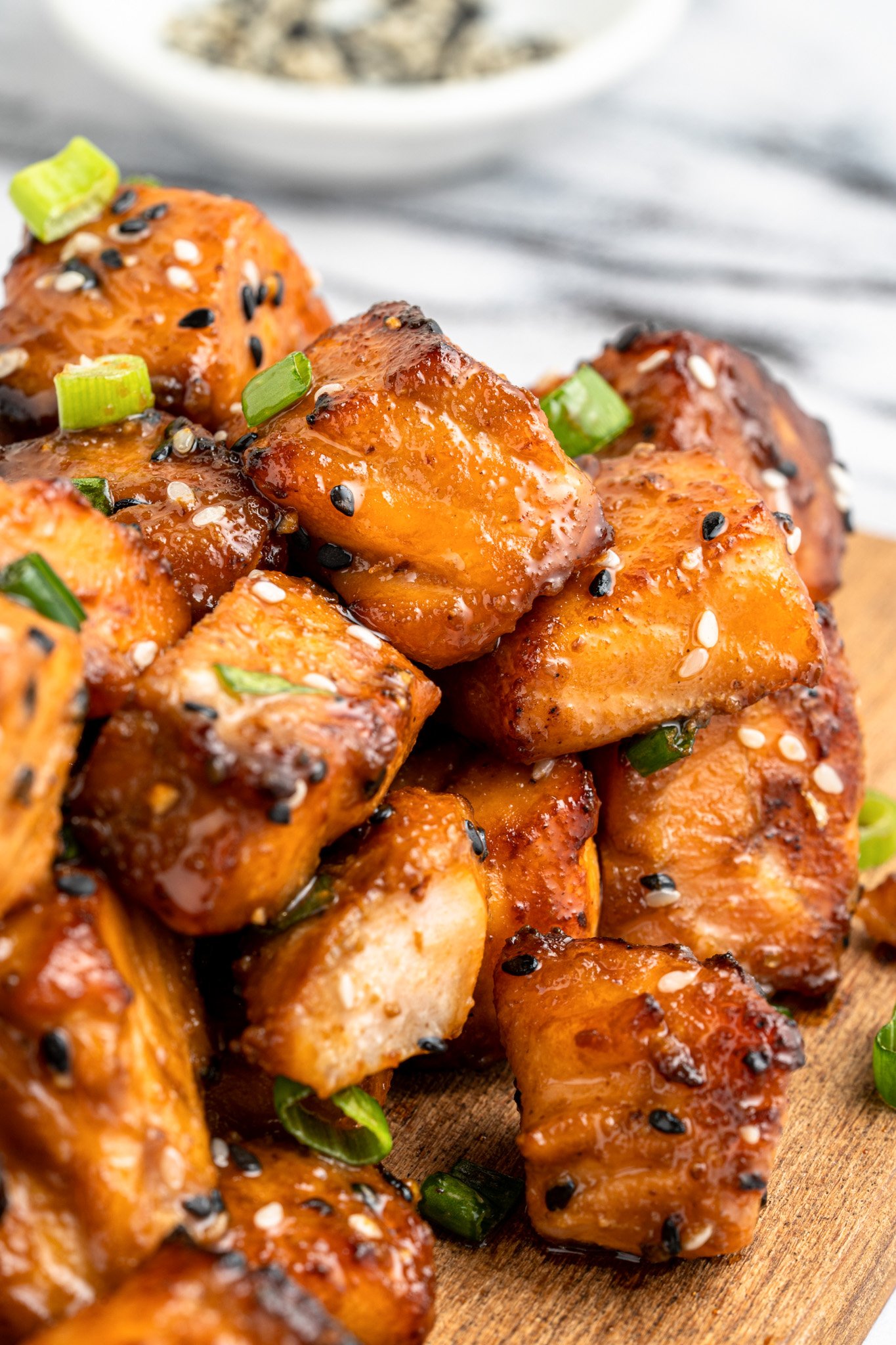 Close-up of glazed salmon cubes garnished with sesame seeds and green onions on a wooden board. A small bowl of sesame seeds is blurred in the background.