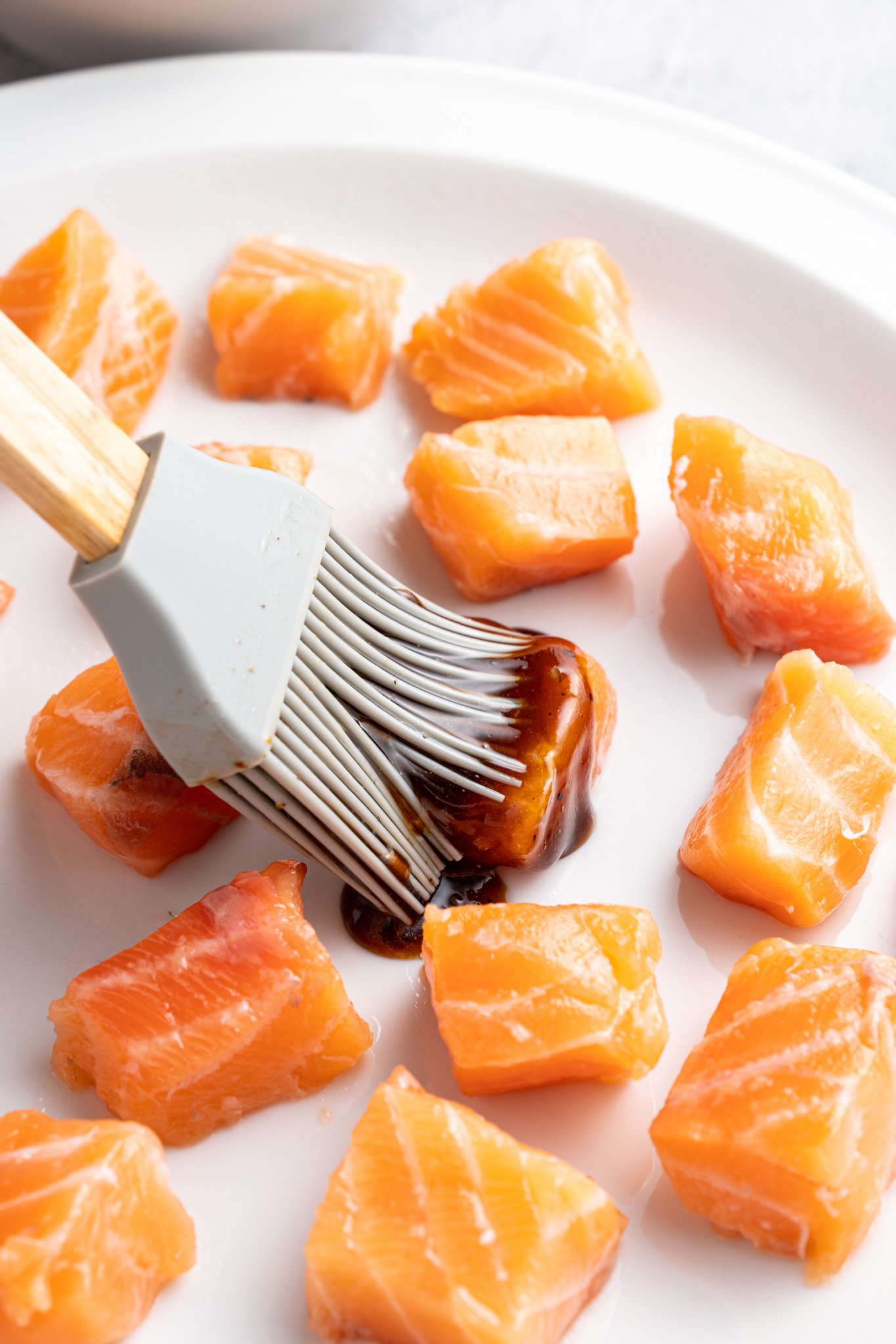 A silicone brush applies a dark marinade to raw salmon cubes on a white plate. The salmon pieces are evenly spaced and glistening, suggesting preparation for cooking. The background is softly blurred.