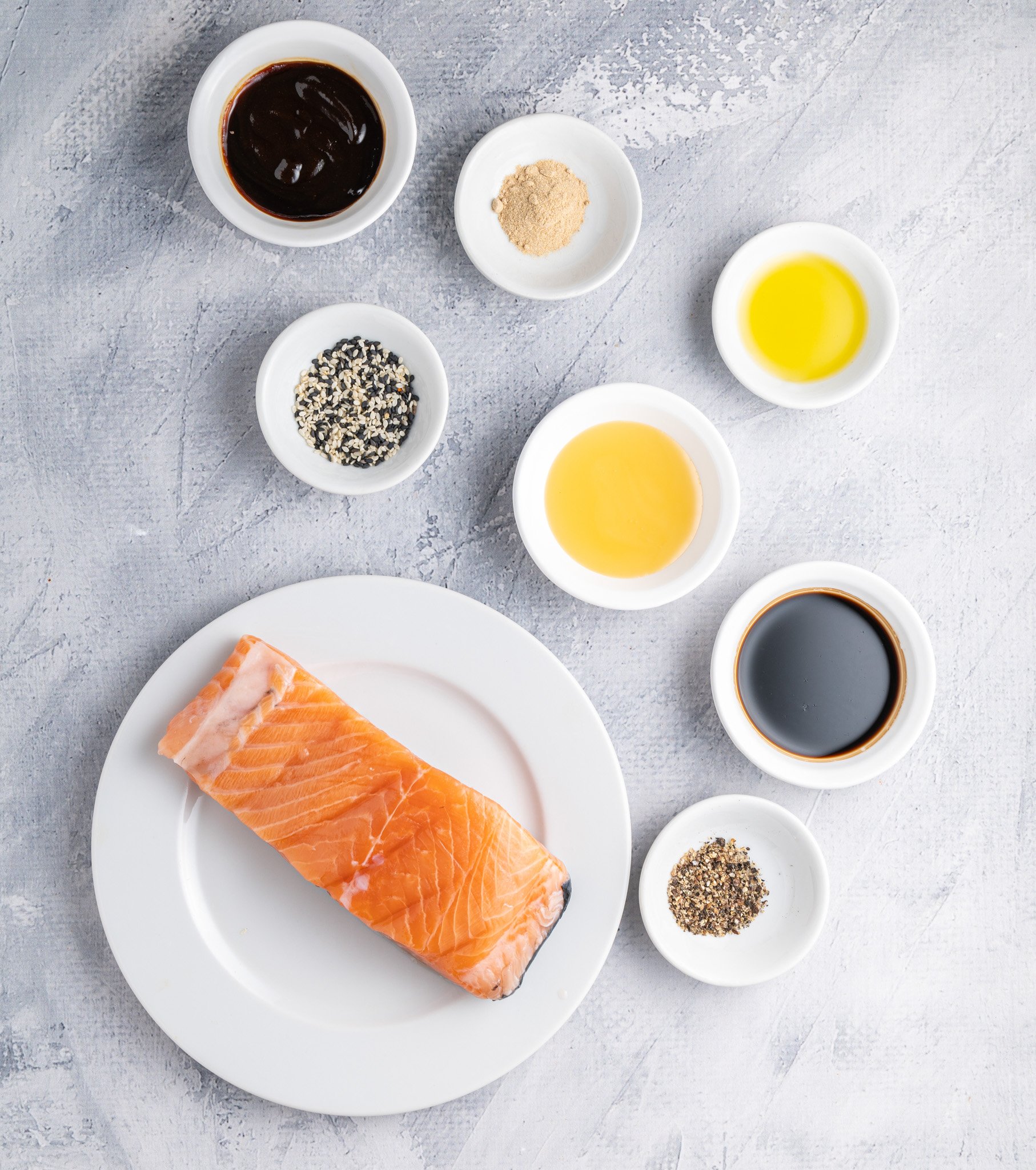 A salmon fillet on a white plate surrounded by small bowls containing various ingredients such as sauce, sesame seeds, oil, garlic powder, and pepper, arranged on a textured gray surface.