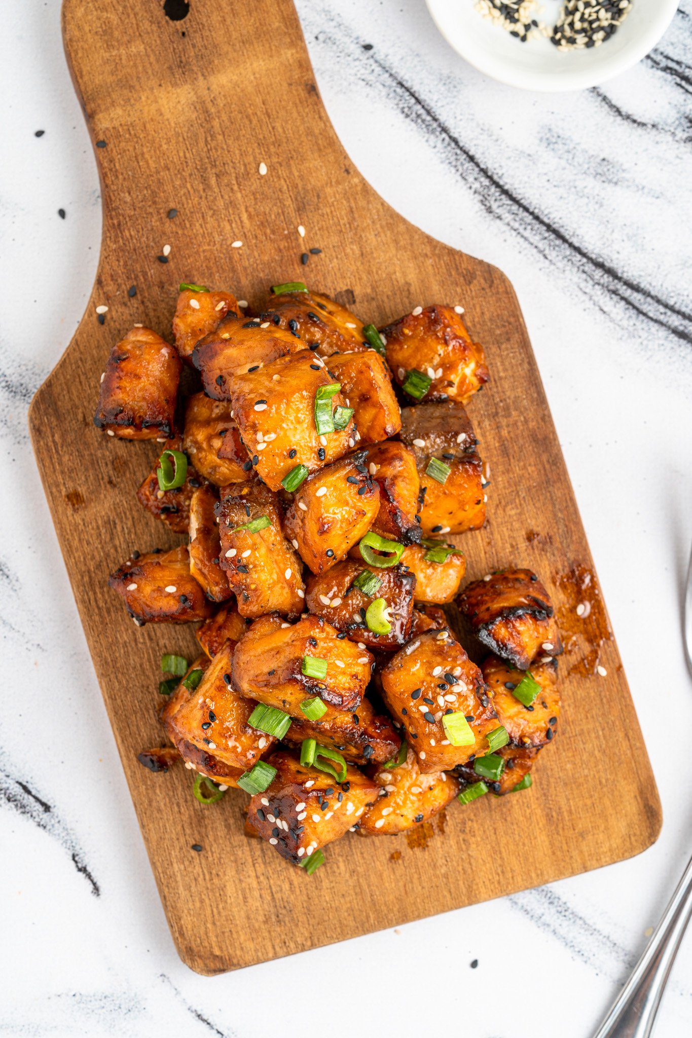 air fryer salmon bites glazed in a brown sauce, garnished with chopped green onions and sesame seeds, served on a wooden cutting board. A small dish with sesame seeds is in the background, set on a marble surface.