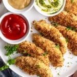Crispy baked chicken fingers on a white plate, golden brown and ready to serve with three dipping sauces.