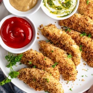 Crispy baked chicken fingers on a white plate, golden brown and ready to serve with three dipping sauces.