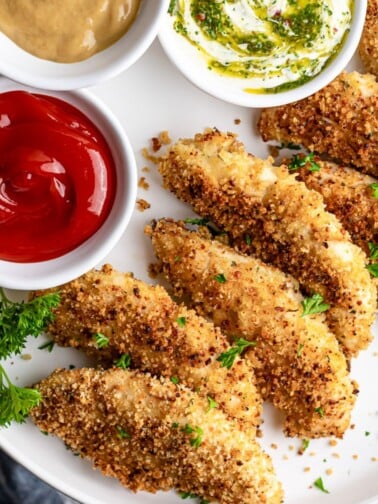 Crispy baked chicken fingers on a white plate, golden brown and ready to serve with three dipping sauces.