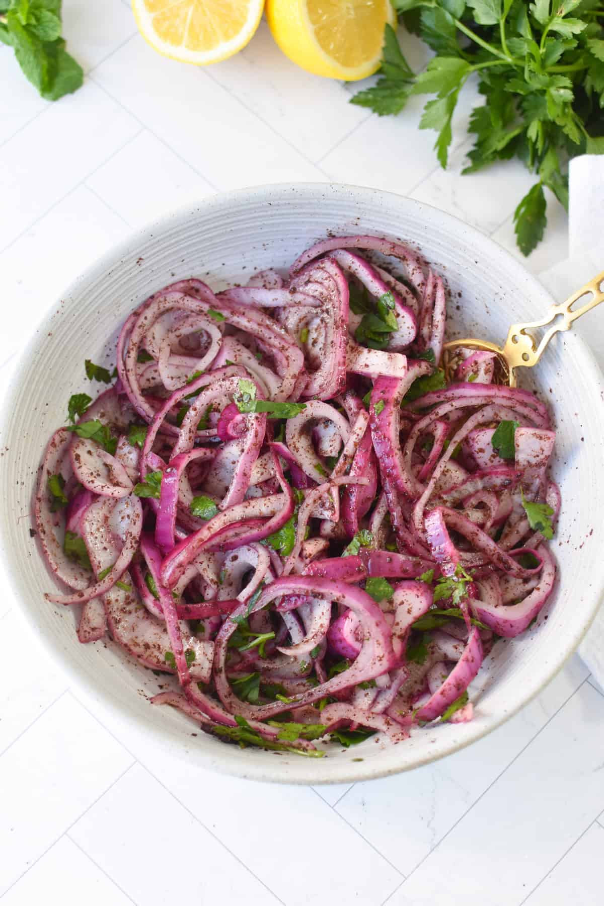 Sumac onions ingredients including red onion, sumac, fresh parsley, lemon, red wine vinegar and olive oil in a white bowl